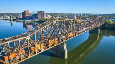 Altın saat boyunca Cincinnati 'deki Ohio Nehri üzerindeki paslı çelik bir köprünün havadan görüntüsü. Aşağıdan geçen bir tren, canlı bir şehir zeminiyle şehir ve sanayi bölgelerini vurguluyor.