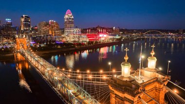 Cincinnati 'nin mavi saatindeki gökyüzü manzarasında Ohio Nehri üzerindeki aydınlatılmış John A. Roebling Askı Köprüsü canlı bir şehrin ışıklarını ve enerjisini yansıtıyor..