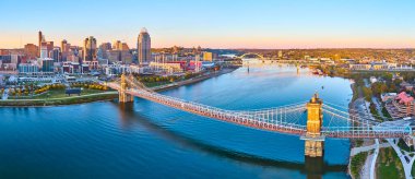 An aerial view of Cincinnati showcases the iconic John A Roebling Suspension Bridge during golden hour. The city skyline and Ohio River create a stunning blend of urban vibrancy and natural beauty. clipart