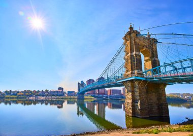 Ohio Nehri üzerindeki John A Roebling Askı Köprüsü 'nün zarafetini yakalayan bu panoramik manzara, modern Cincinnati siluetine karşı olan tarihi yapıyı berrak mavi bir gökyüzü altında vurguluyor.