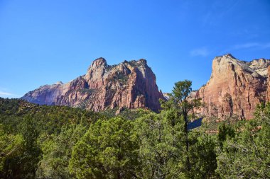 Zion Ulusal Park Sarayı 'nın canlı güzelliklerine dalın. Kızıl kumtaşı tepeleri, yemyeşil ağaçlar ve açık mavi gökyüzü sizi bu doğal harikayı keşfetmeye davet ediyor..