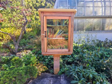 Charming free library nestled in a lush garden invites community sharing and reading in Cincinnati Ohio. Surrounded by vibrant greenery and a greenhouse backdrop, it is a serene escape for book lovers clipart