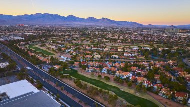 Gün batımında Las Vegas banliyösünün havadan görünüşü canlı yeşil golf sahası ve engebeli dağlara yaslanmış kırmızı kiremitli evler altın saat içinde kentsel büyüme ve yaşam tarzının özünü yakalıyor..