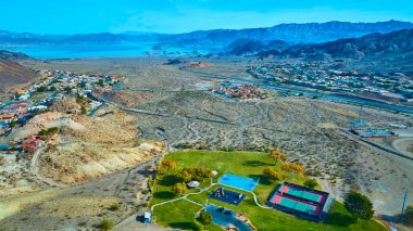 Aerial view of Boulder City in Nevada reveals a lush park oasis with playground and tennis courts, surrounded by desert landscapes, a serene Lake Mead, and majestic mountains under clear blue skies. clipart