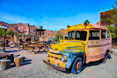 Nelson, Nevada 'nın nostaljik çekiciliğini bu kırsal çöl sahnesiyle keşfedin. Piknik masalarının yanında yıpranmış sarı bir vintage araç ve parlak mavi gökyüzünün altında tuğla bir yapı duruyor..