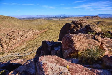Las Vegas, Nevada yakınlarındaki Red Rock Kanyonu 'nun görkemli manzarası. Kırmızımsı engebeli kaya oluşumları ve seyrek bitki örtüsü uzak bir şehir manzarasına karşı çöl manzarasının dingin güzelliğini vurgular..