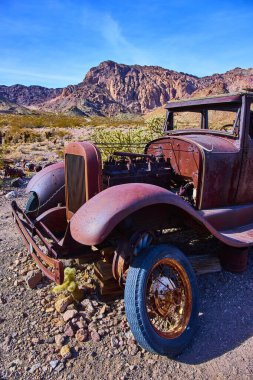Paslı eski araba Nevada 'nın Nelson çölünde dinleniyor. Terk edilmiş nostalji, engebeli dağlar ve seyrek kaktüsler gibi doğayla buluşur. Gün ortası güneşinin altında zamansız bir manzara resmeder..