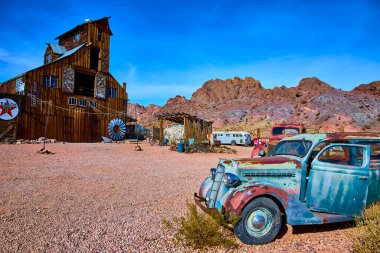 A rusted vintage car rests among desert shrubs in Nelson Nevadas ghost town. A weathered barn with a Texaco sign and classic vehicles evoke nostalgia and the allure of a bygone Americana era. clipart