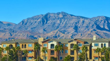 Las Vegas, Nevada yakınlarındaki engebeli dağ sıralarının çarpıcı zeminine karşı kurulmuş, gür palmiyeler ve parlak mavi gökyüzü altında çerçevelenmiş zarif çok katlı konutların havadan görünüşü.