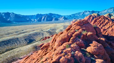 Nevada 'daki Kızıl Kaya Kanyonu' nun hava manzarasında açık mavi gökyüzü altında parlak kırmızı kumtaşı oluşumları ve görkemli dağlar görülüyor. Amerika 'nın güneybatısının doğal güzelliğini ve macerasını tecrübe edin.