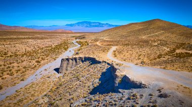 Nevada 'daki Charleston Dağı yakınlarındaki engebeli bir çöl arazisinin dolambaçlı toprak yolunun hava görüntüsü. Nadir bitki örtüsü ve uzak dağlar açık mavi gökyüzü altında macera ve keşif çağrıştırıyor..