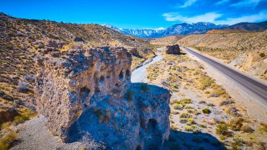 Nevada Çölü 'nün engebeli kaya oluşumları ve Charleston Dağı' nın karla kaplı olduğu uzak bir yol manzarası. Uçsuz bucaksız mavi gökyüzünün altında baş döndürücü bir yalnızlık ve macera karışımı belirir..