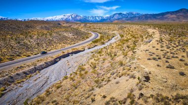 Nevada çölünde dönen siyah bir arabanın hava görüntüsü, Joshua ağaçları ve karla kaplı Charleston Dağı. Seyahat, macera ve açık yolların cazibesi için mükemmel..