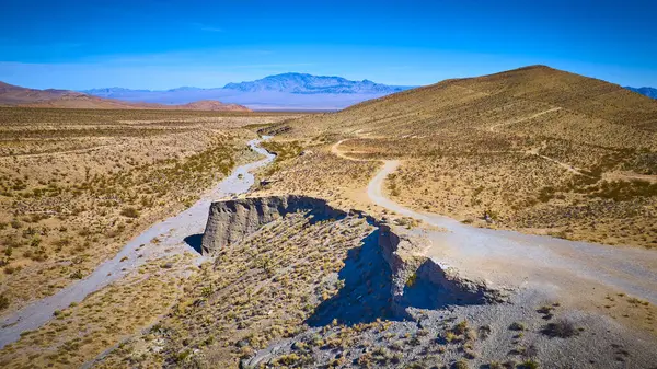 Nevada 'daki Charleston Dağı yakınlarındaki engebeli bir çöl arazisinin dolambaçlı toprak yolunun hava görüntüsü. Nadir bitki örtüsü ve uzak dağlar açık mavi gökyüzü altında macera ve keşif çağrıştırıyor..
