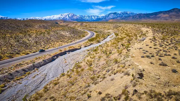 Nevada çölünde dönen siyah bir arabanın hava görüntüsü, Joshua ağaçları ve karla kaplı Charleston Dağı. Seyahat, macera ve açık yolların cazibesi için mükemmel..