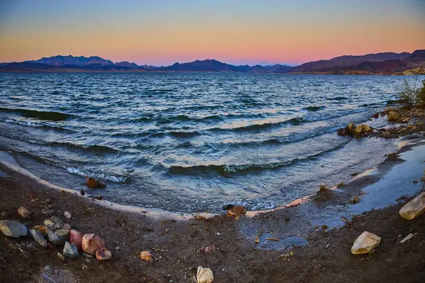 Nevada 'daki Mead Gölü' nde altın saat kayalık bir sahil şeridinde sakin dalgaların görüntüsünü yakalıyor. Gün batımı, uzak dağların çerçevelediği gökyüzünü ve suyu yumuşak renklerle boyar. Temalar için mükemmel