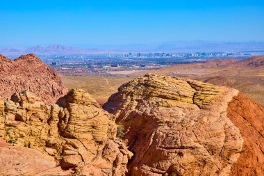 Calico Tank Yolu 'ndan göz kamaştırıcı manzara: Kırmızı Kaya Kanyonu' nun canlı kırmızı ve sarı kumtaşı oluşumları parlak mavi gökyüzünün altında Las Vegas silueti ile buluşuyor. Doğa ve şehir mükemmel bir uyum içinde..