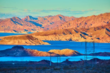 Altın saat, Meads Gölü 'nün üzerinde dingin, mavi suları ve çöl dağlarını aydınlatıyor. Elektrik hatları, doğayı Boulder City, Nevada yakınlarındaki sanayiyle harmanlayarak araziye yayılıyor..