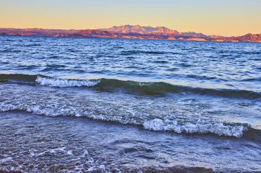 Altın güneş ışığı Nevada, Boulder City 'deki Mead Gölü' nün sakin kıyısına vurur. Dalgalar yavaşça kumu kaplar uzak dağlar gibi gün batımına karşı siluet, sakin doğayı yakalar.