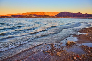 Nevada 'daki Mead Gölü' nde altın saat. Gün batımı dağları sıcacık bir ışıltıyla yıkarken yumuşak dalgalar kayalık sahili öper. Huzurlu bir kaçış doğaya huzur dolu bir kucaklama.