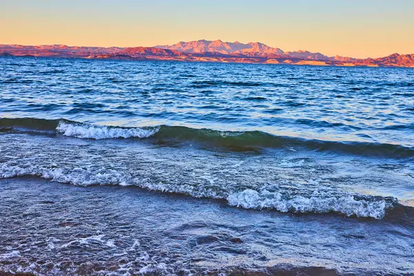 Altın güneş ışığı Nevada, Boulder City 'deki Mead Gölü' nün sakin kıyısına vurur. Dalgalar yavaşça kumu kaplar uzak dağlar gibi gün batımına karşı siluet, sakin doğayı yakalar.