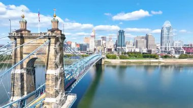Aerial Fly Through Cincinnatis nehir cephesinde ikonik asma köprüsü, canlı ufuk çizgisi, ve sakin Ohio Nehri, kentsel gelişimin ve doğal güzelliğin ahengini temsil ediyor..