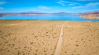 Mead Gölü yakınlarındaki Nevadas Çölü 'nün virajlı toprak yolundaki yalnız bir aracın geniş hava görüntüsü, uzak dağlar tarafından çerçevelenmiş. Macera, keşif ve yalnızlık temaları için mükemmel..