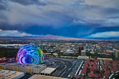 Las Vegas 'ın havadan görünüşü girdap gibi neon desenlerle The Sphere' i gösteriyor. Öğleden sonra gökyüzü fırtınayı ima ediyor, bu canlı şehir manzarası için dramatik bir zemin oluşturuyor..