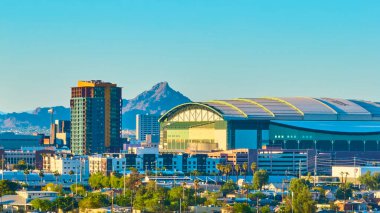 Phoenix şehir merkezinin altın saat hava manzarasında modern yüksek binalar Chase Field Stadyumu ve Camelback Dağı 'nın palmiye ağaçları ve çöl yeşillikleri ile canlı Arizona şehir manzarasını vurguluyor.