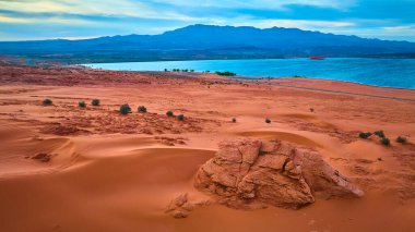 Utah 'taki Sand Hollow Gölü' nün mavi sularının yanında kızıl kum tepelerinin ve engebeli kaya oluşumlarının havadan görünüşü. Uzak dağlar ve dramatik gökyüzü Amerika 'nın güneybatısının güzelliğini vurguluyor.