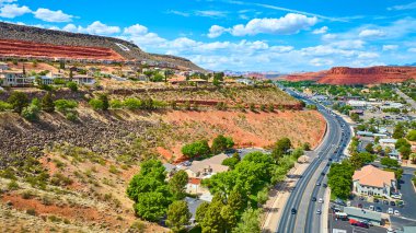 St. George Utah 'taki dramatik kırmızı kaya tepesindeki modern evlerin havadan görünüşü aşağıdaki dolambaçlı işlek bir yol ve beyaz bulutlu parlak mavi bir gökyüzünün altında uzanan banliyö şehri.