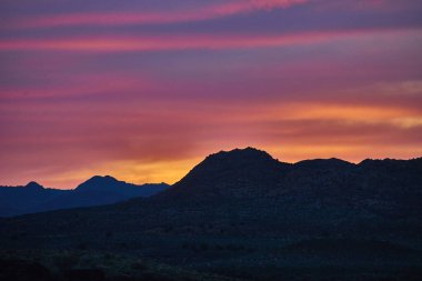 Günbatımının canlı renkleri, Utah Çölü yakınlarındaki engebeli dağ siluetleri üzerinde gökyüzünü aydınlatıyor. Huzurlu ve ilham verici bir Amerikan Güneybatı manzarası yaratıyor..
