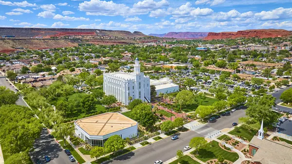 St. George Utah 'ın havadan görünüşü, açık mavi gökyüzü altında yemyeşil banliyö evleri ve manzaralı kızıl kaya birikimleriyle çevrili çarpıcı beyaz St. George Utah Tapınağı' nı gösteriyor.