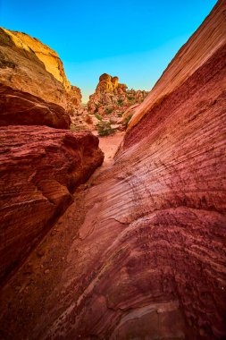Canlı kırmızı ve turuncu kumtaşı katmanları Utah Kasırgası 'nda Babil Kemeri boyunca kıvrılıyor. Açık mavi gökyüzü altında engebeli kaya oluşumları ve seyrek çöl bitkileri bu ikonik manzaranın güzelliğini vurguluyor.