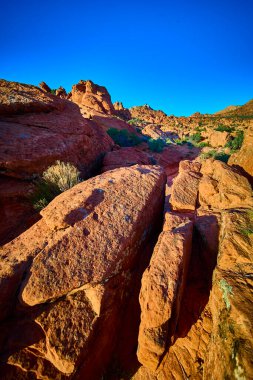Güneşli kırmızı kumtaşı kayaları ve engebeli kaya oluşumları Babil Arch Utah yakınlarındaki çöl arazisine hükmediyor. Parlak mavi gökyüzünün altında dayanıklı çöl bitkileri yeşil dokunuşlar ekliyor..