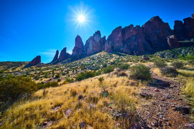 Güneşin öptüğü kaya oluşumları, Arizona 'daki Kayıp Hollandalı Eyalet Parkı' nda parlak mavi bir gökyüzü altında altın çöl çimlerinin üzerinde yükseliyor ve Amerika 'nın güneybatısının vahşi güzelliğini ve macerasını yakalıyor..