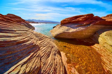 Çarpıcı kırmızı kumtaşı oluşumları Utah Kasırgası yakınlarındaki Sand Hollow Eyalet Parkı 'nda güneşli bir havuz oluşturuyor. Uzak dağlara karşı çarpıcı dokuları ve berrak suları ve uçsuz bucaksız mavi gökyüzü var..