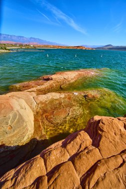 Güneşli kırmızı kumtaşı kayaları Utah 'taki Sand Hollow State Park Gölü' nün berrak turkuaz sularını çevreleyen açık hava maceraları ve seyahatler için mükemmel mavi gökyüzü altındaki uzak dağlarla çevreler.