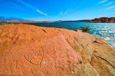 Utah 'taki Sand Hollow Eyalet Parkı' nda güneş ışığı altındaki kırmızı kumtaşı, doğada sevgi ve macera yakalayan, parıldayan mavi göl ve çöl manzarasına bakan oyulmuş bir kalp ve baş harfler barındırır..
