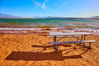 Utah 'ta kumlu bir gölün kumlu sahilinde bir metal piknik masası. Mavi suları ve berrak bir gökyüzünün altındaki uzak dağları var. Manzaralı bir ortamda rahatlama ve açık hava keyfine davet ediyor..