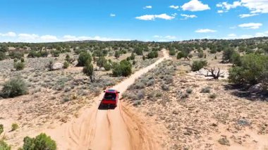 Arizona Çölü 'nde, Mermer Kanyon ve Vermilion Kayalıkları' nın yakınlarındaki uzun toprak yolda ilerleyen kırmızı bir kamyonun üzerinden uçan hava aracı engebeli bir arazi ve güneşli bir bitki örtüsünün altında seyrek bir bitki örtüsüyle çevrili.