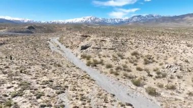 Havadan Uçuşan İlerleme: Nevada Dağı 'nın engebeli güzelliğini keşfedin. Kurak arazi ve açık mavi gökyüzünün altındaki dolambaçlı toprak yol ile karla kaplı zirveler. Doğa için mükemmel.