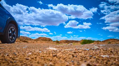 Mavi araba ve engebeli lastik kayalık çöl zemininde canlı bir gökyüzünün altında dağınık beyaz bulutlar ve uzak tepeler macera ve keşif hissi yaratıyor.