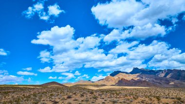 Uçsuz bucaksız çöl arazisi engebeli dağ sırtlarıyla buluşur. Beyaz bulutlarla dolu dramatik bir gökyüzü ve parlak gün ortası ışığı altında.