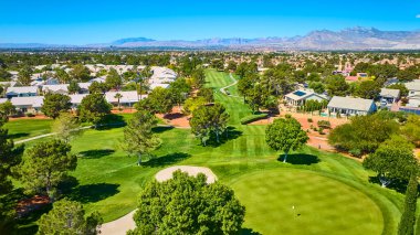 Canlı bir golf sahasının geniş hava manzarası banliyö evleriyle dolu ve berrak mavi gökyüzünün altındaki dağ sıraları tarafından çerçevelenmiş.