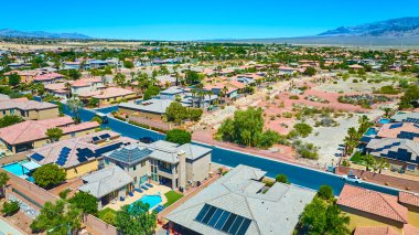 Açık mavi gökyüzü altında uzak dağların zeminine kurulmuş havuzlu, güneşli ve çöl arazili banliyö evlerinin parlak hava manzarası