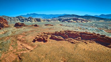Uçsuz bucaksız kızıl kaya oluşumları engebeli çöl arazileri ve açık mavi gökyüzünün altındaki uzak dağlar.