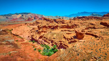 Canlı kırmızı kaya oluşumlarının geniş hava manzarası engebeli kanyonlar ve açık mavi gökyüzünün altındaki seyrek çöl bitkileri.