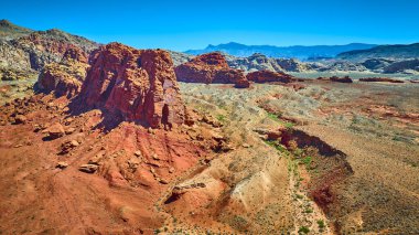 Parlak kırmızı kumtaşı uçurumları, engebeli çöl arazisinin üzerinde, uzak sıradağları olan açık mavi gökyüzünün altında.