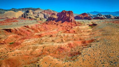 Canlı kırmızı kayalar, engebeli çöl arazisinin üzerinde, açık mavi gökyüzünün altında, dağınık kurak bitki örtüsüyle yükseliyor.